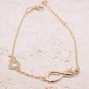 Gold bracelet with heart and infinity design on a beige surface