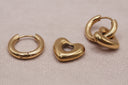 Three gold-toned earrings displayed on a light beige textured surface: two small hoop earrings and one polished heart-shaped hoop with a center cutout.