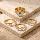 Amore Heart Cluster Stackable Ring Set- Four gold rings displayed on beige stone blocks. The set includes a stack of three rings with a four-leaf clover motif paved with clear stones, a ring with a small heart-shaped clear-stone setting, a bow-shaped clear-stone setting, and another heart-shaped clear-stone setting.