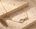 A delicate gold chain necklace featuring an infinity symbol pendant with a small clear stone, and a small heart-shaped pendant, displayed on textured beige stone blocks.