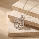 A silver tree of life pendant necklace with a circular frame, displayed on stacked beige stone tiles.