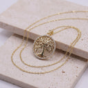 A gold tree of life pendant necklace with a circular bezel-set crystal border, displayed on a textured beige stone slab.