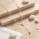 A delicate silver Y-necklace with three elongated teardrop charms, displayed on stacked beige travertine stones with small white decorative pebbles.