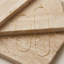 Ivy Barely There Box Chain Necklace. A pair of delicate gold chain earrings displayed on stacked beige travertine stone tiles.