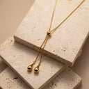 Aurelia Adjustable Lariat Necklace. A gold lariat-style necklace with two teardrop-shaped gold pendants, displayed on stacked beige travertine stone blocks.