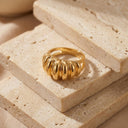 Sculpt Statement Ring- A gold ring with a textured, ribbed design, displayed on stacked beige travertine stone tiles.