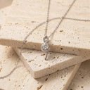 Elia Moissanite Twist Necklace – 1ct VVS | 925 Sterling Silver. A silver necklace with a unique loop pendant set with a round clear gemstone, resting on stacked beige travertine stone tiles.