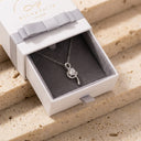 Elia Moissanite Twist Necklace – 1ct VVS | 925 Sterling Silver. A silver-toned flower pendant necklace with a clear center stone, displayed inside an open white Ella & Faith London gift box with a gray ribbon bow, set on a beige stone surface.
