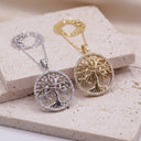 Two  tree-of-life pendant necklaces displayed on beige stone blocks, one in silver with a silver chain and one in gold with a gold chain. The pendants feature detailed tree motifs with small clear stones around the edges.