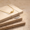 Two delicate leaf-shaped PENDANTS, one gold and one silver, draped over stacked beige stone blocks on a matching stone surface.