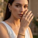 A close-up lifestyle shot of a woman wearing Elara Link Bracelet on her wrist, a gold hoop earring, and a gold band ring with clear stones, set against a warm, softly blurred outdoor background.