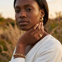 A woman in a white top wearing Elara Link Bracelet, a gold ring, and a gold hoop earring, posing outdoors with a blurred natural background.