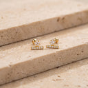 A pair of Star & Bar CZ Cartilage Stud with a small clear gemstone cluster on top of a horizontal bar set with tiny clear stones, displayed on a beige stone surface.