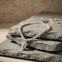 A silver beaded bracelet with an acorn pendant, styled on stacked dark gray slate stones against a textured beige background.