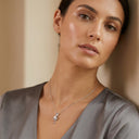 Elia Moissanite Twist Necklace – 1ct VVS | 925 Sterling Silver. A close-up of a woman wearing a delicate silver necklace with a small sculptural pendant, styled with a gray satin top against a warm beige background.
