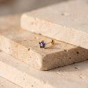 Sapphire Flora Flower Piercing Stud- A small gold stud earring with a cluster of deep blue gemstones, resting on stacked beige travertine stone blocks.