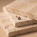 Sapphire Trio Piercing Stud - A small gold body jewelry piece with blue gemstones rests on stacked beige travertine stone slabs against a warm beige background.