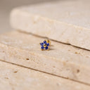 Sapphire Flora Flower Piercing Stud - A small gold flower-shaped stud earring with deep blue gemstones, resting on a textured beige stone surface.