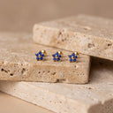 Sapphire Flora Flower Piercing Stud- Three small gold flower-shaped stud earrings with blue gemstones, displayed on a beige stone slab against a warm beige background.