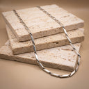 Sculpted Link Necklace. A silver-toned chain necklace with elongated, polished links draped over stacked rectangular stone tiles on a warm beige background.