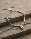 A silver beaded slider bracelet with a small acorn charm, displayed on layered grey stone slabs.