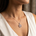 A close-up of a woman's neck and chest wearing a delicate silver chain necklace with a circular tree-of-life pendant, styled with a white sleeveless top.