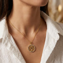 A close-up of a model wearing a delicate gold tree of life pendant necklace with a small halo border, styled with a white textured blouse.
