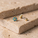 Himari Sunflower Piercing Stud- A pair of small flower-shaped stud earrings with turquoise blue petals and gold centers, shown with one earring turned to display the post and backing on a beige stone surface.