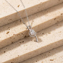 Elia Moissanite Twist Necklace – 1ct VVS | 925 Sterling Silver. A silver necklace with a unique pendant featuring a round clear gemstone surrounded by small clear stones, resting on beige textured stone blocks.