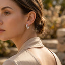 Ayra Twist Hoop Earrings -A woman in a beige top wearing a small silver huggie hoop earring with a textured, twisted design, photographed in a warm, softly blurred outdoor setting.