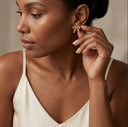 A close-up lifestyle shot of a woman wearing a small gold bow-shaped stud earring, touching it with her finger. She is wearing a cream-colored camisole against a soft, warm beige background.