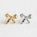 Two small bow-shaped stud earrings, one in polished gold and one in polished silver, displayed side-by-side on a light textured white surface.