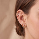 Close-up of a model's ear showcasing Flora Vine CZ Cartilage Stud alongside other gold ear piercings, against a soft beige background.
