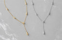 Two Y-necklaces with tapered cone charms, one in gold and one in silver, displayed on a light gray marble surface.
