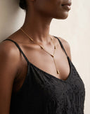 A close-up of a model wearing a delicate gold Y-necklace with a tapered drop pendant, paired with a black lace camisole top against a neutral beige background.