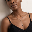 A close-up of a model wearing a delicate silver Y-shaped necklace with a tapered drop pendant, paired with a black spaghetti-strap top against a neutral beige wall.