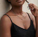 A close-up lifestyle shot of a model wearing a delicate silver Y-necklace with tapered geometric accents, paired with a black lace camisole against a neutral textured wall.