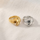 Two twisted wrap-style rings, one in polished yellow gold and one in polished silver, displayed side-by-side on a textured off-white fabric background.