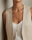 A close-up of a model wearing a delicate gold lariat necklace with two dangling bead chains over a white top and a beige linen vest.