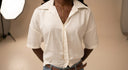 A person wearing a short-sleeve white button-up shirt with a relaxed fit, paired with blue jeans and a gold necklace, standing in a studio setting.