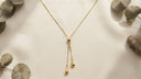 A delicate gold Y-necklace with two teardrop pendants, styled on a warm off-white surface with dried eucalyptus leaves.