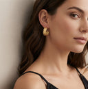 A close-up of a woman wearing a textured gold crescent-shaped hoop earring, leaning against a neutral beige wall.
