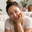 A woman with her hair in a bun rests her head on her hand, wearing a delicate gold chain bracelet and matching ring. She is wearing a light beige ribbed t-shirt in a cozy indoor setting with soft-focus plants in the background.
