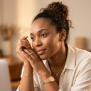 A woman with her hair in a bun wearing a white linen shirt and gold jewelry, including a gold watch, delicate necklace, and rings, resting her chin on her hands in a warm, softly lit indoor setting.