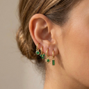 Close-up of an ear wearing three gold hoop earrings with green gemstones on a neutral background.