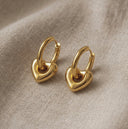 A pair of gold hoop earrings with a polished heart-shaped drop on each earring, displayed on a textured beige linen fabric background.