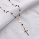 A delicate gold chain necklace with small black beads, a round gold medallion, and a cross pendant, displayed on a soft white fabric background.