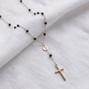 A delicate gold chain necklace with small black beads, a round gold medallion, and a cross pendant, displayed on a textured white fabric background.