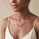 A close-up of a woman wearing layered necklaces, featuring a delicate gold chain with small black beads and a cross pendant, layered over a thin gold chain.