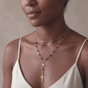 Gold Christian cross necklace layered with delicate chain- A close-up of a woman wearing a delicate gold necklace with a cross pendant and small black beads, layered over a thin gold chain necklace, paired with a cream-colored camisole top.
